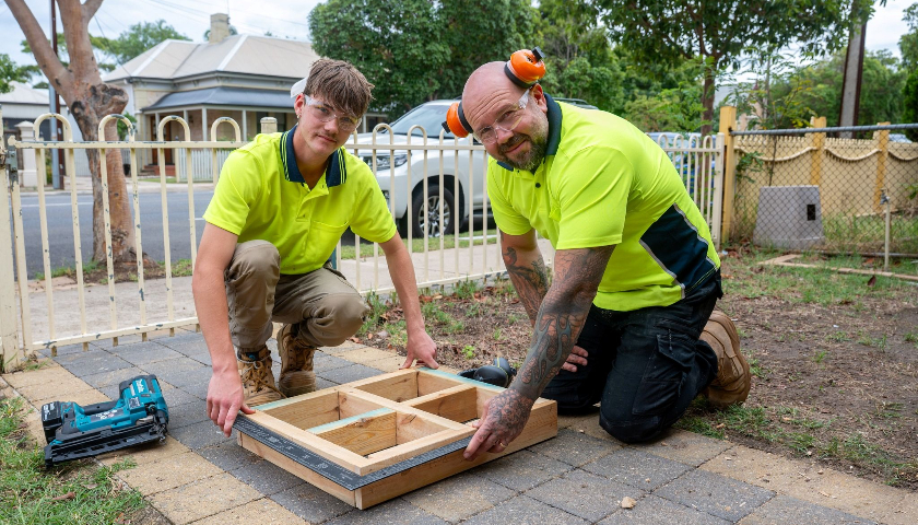 A younger man and an older man wearing yellow high‑visibility tops smile at the camera while working with timber.