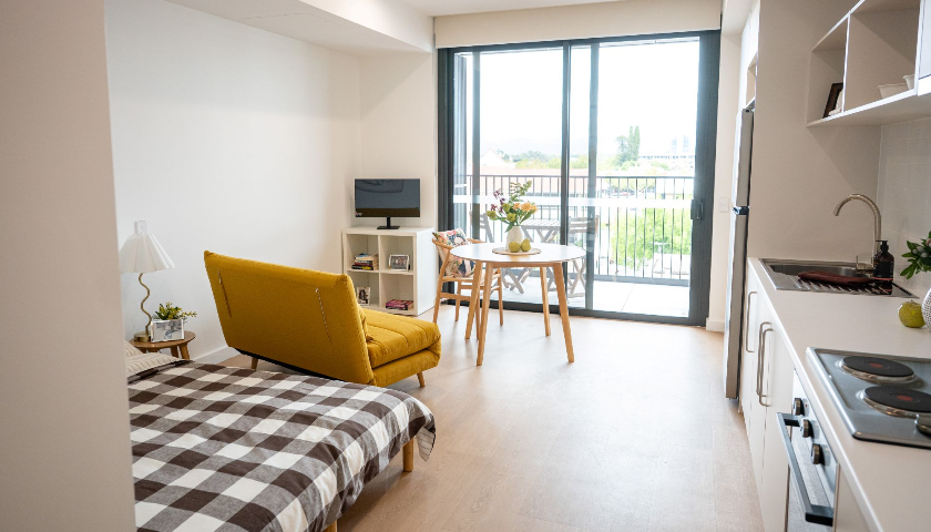 An open-plan room with a bed, yellow chair, kitchen, TV, and glass sliding door.